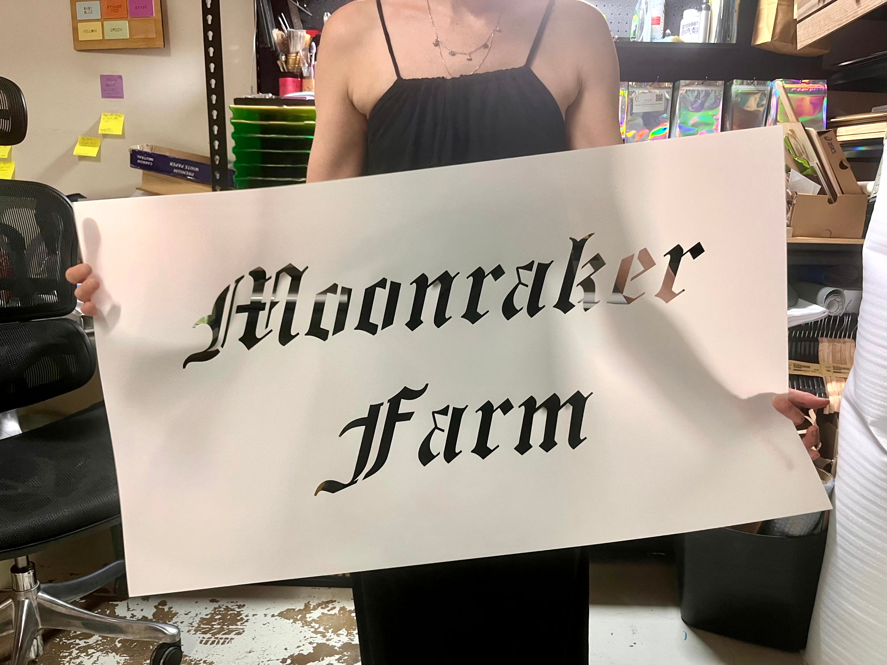 A person holding a large white stencil with the words "Moonraker Farm" cut out in a blackletter gothic font.