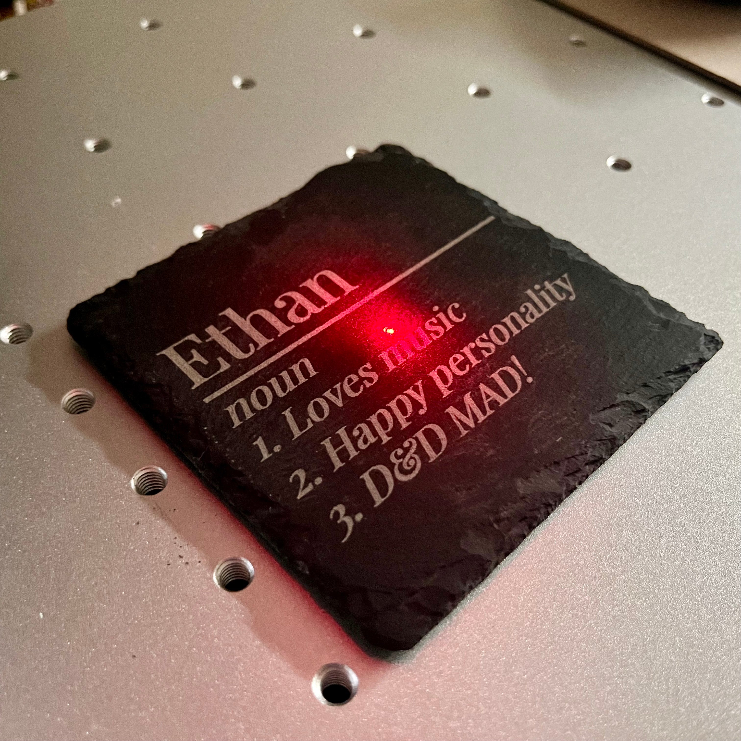 A red laser point engraving text onto a square black slate coaster placed on a silver metal laser bed.