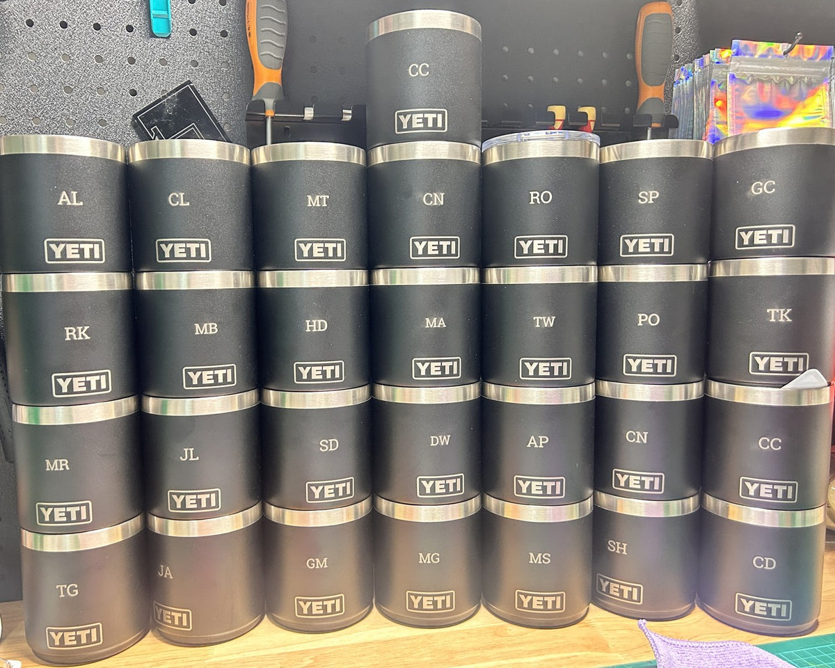 Twenty-eight black YETI Rambler tumblers stacked in rows, each laser engraved with a different set of initials